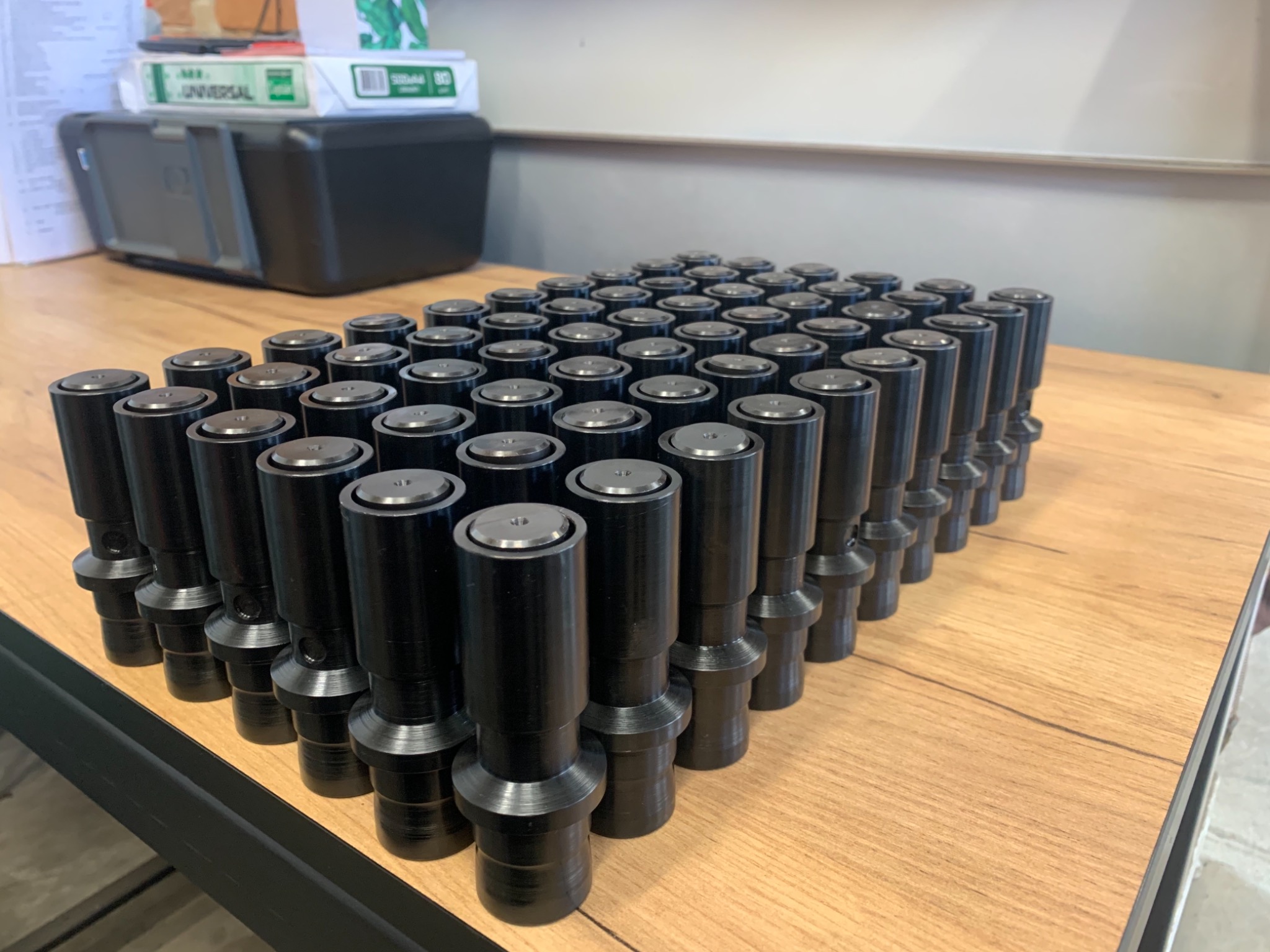 Partia czarnych, metalowych elementów cylindrycznych z kołnierzami ułożonych na drewnianym blacie, w tle drukarka i kartony papieru.