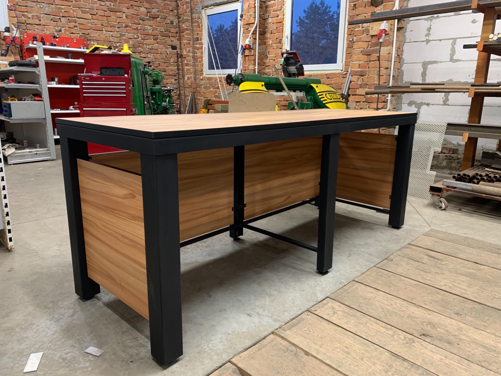 Stół spawalniczy z blatem z jasnego drewna i czarną metalową konstrukcją, w tle warsztat z ceglaną ścianą i maszynami.