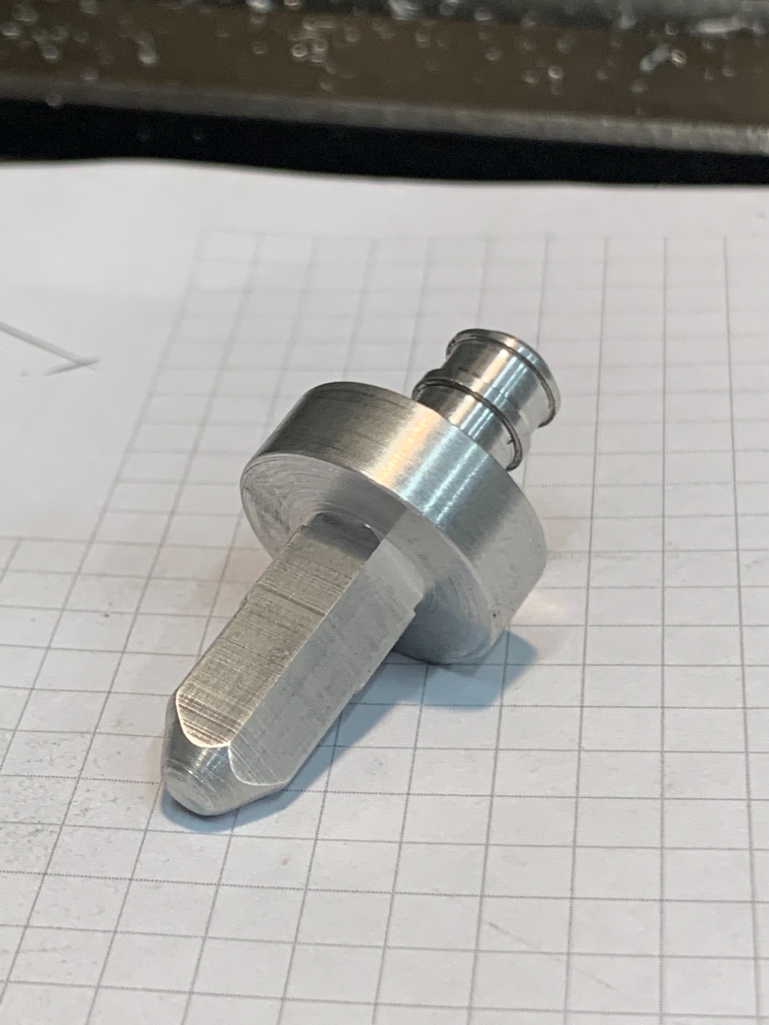 Metalowy element obrobiony na tokarce, leżący na kartce w kratkę. Widoczne ślady obróbki skrawaniem.