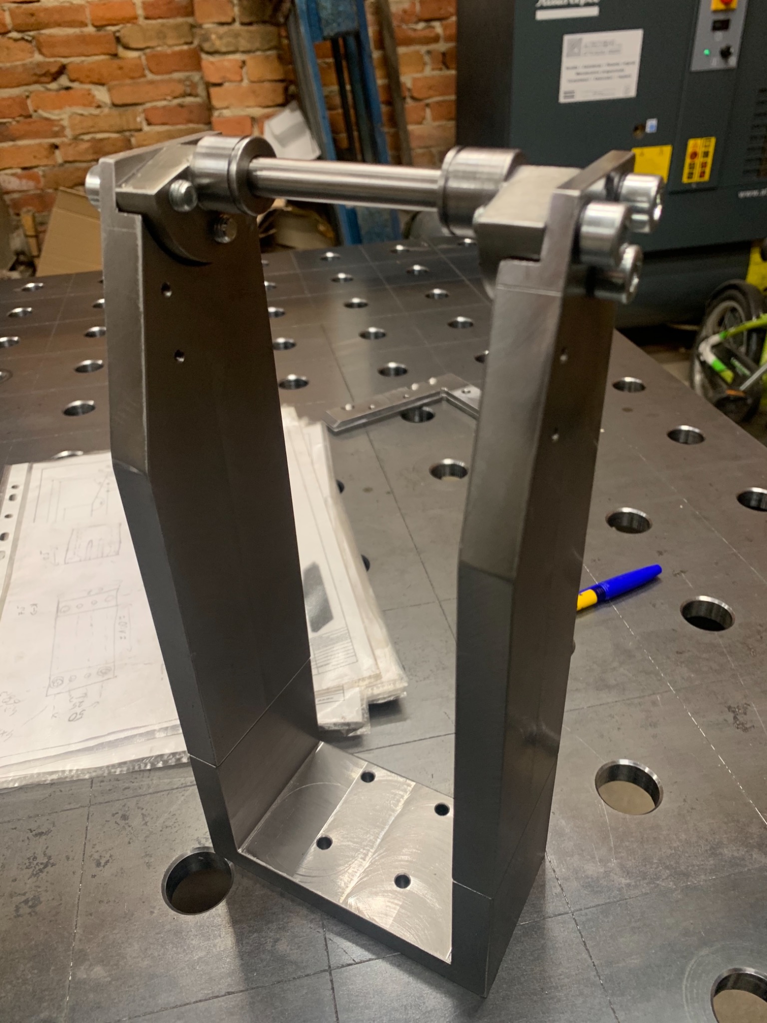 Metalowa konstrukcja ramowa z poprzeczką, złożona na stole spawalniczym z otworami, obok leżą rysunki techniczne i niebieski długopis.