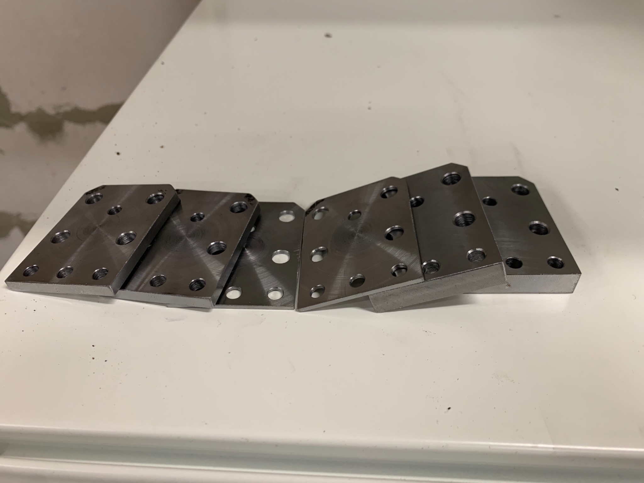 Sześć metalowych, prostokątnych płytek z otworami ułożonych na białym blacie. Płytki mają surowe wykończenie i widoczne ślady obróbki.