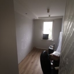 Krystian koseski - Pokój w Radzyminie w trakcie przygotowania do malowania: ściany po szpachlowaniu, podłoga zabezpieczona, widoczny kufer z narzędziami i krzesło biurowe.