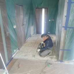 Krystian koseski - Remont korytarza: mężczyzna w okularach i rękawicach, na kolanach, usuwa starą posadzkę z użyciem młota udarowego, ściany i drzwi zabezpieczone folią ochronną.