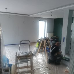 Krystian koseski - Mężczyzna w kombinezonie maluje wałkiem szarą ścianę w pomieszczeniu zabezpieczonym folią, widoczne drabiny, narzędzia i puszki z farbą.