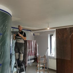 Krystian koseski - Mężczyzna w kombinezonie roboczym stoi na aluminiowej drabinie z dwiema szpachelkami w rękach, przygotowując się do szpachlowania sufitu w pomieszczeniu zabezpieczonym folią malarską.