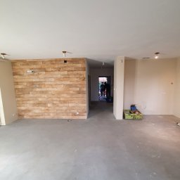 Krystian koseski - Wnętrze w trakcie prac wykończeniowych: ściana z dekoracyjnej cegły, surowa podłoga, przygotowane ściany do malowania, widoczne puszki elektryczne i punkty oświetleniowe.