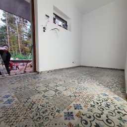 Krystian koseski - Wnętrze w trakcie prac wykończeniowych: świeżo ułożona, wzorzysta podłoga z płytek ceramicznych, białe ściany z zamontowanym oknem i wystającymi przewodami elektrycznymi, widoczny fragment drabiny...