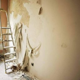 Krystian koseski - Przygotowanie ściany do remontu: zerwana stara tapeta odsłania nierówności, aluminiowa drabina oparta o ścianę, listwa przypodłogowa leży na podłodze wśród resztek tapety.
