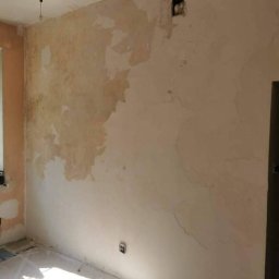 Krystian koseski - Ściana w trakcie przygotowania do malowania, z widocznymi fragmentami starej farby i świeżo nałożonej warstwy wyrównującej, zabezpieczona folią podłoga, w tle żyrandol.