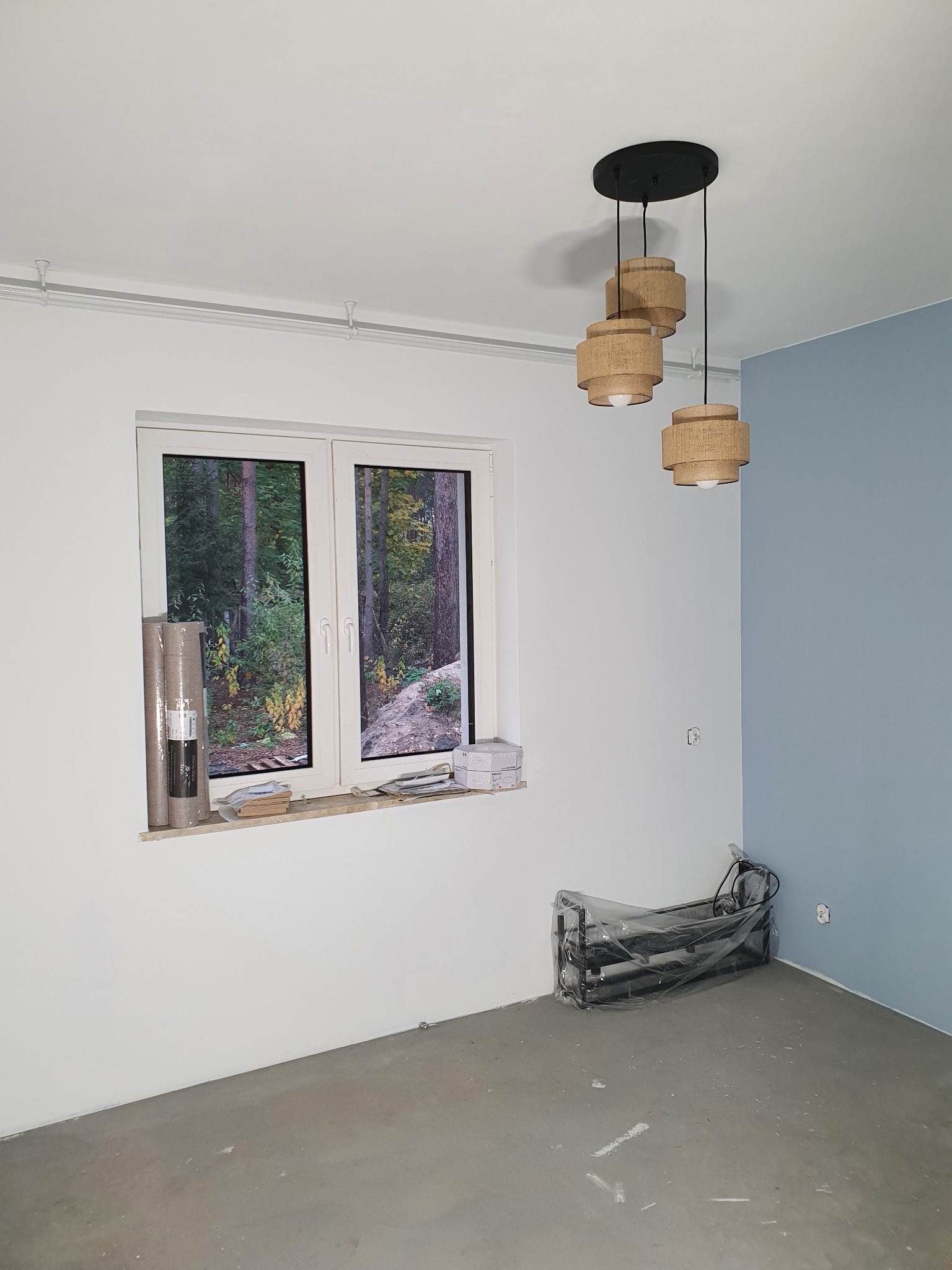 Pokój w trakcie malowania: ściana pomalowana częściowo na błękitno, widoczny nowy parkiet, lampa z trzema abażurami, okno z widokiem na las, rolki tapety przy oknie i sprzęt budowlany w folii.