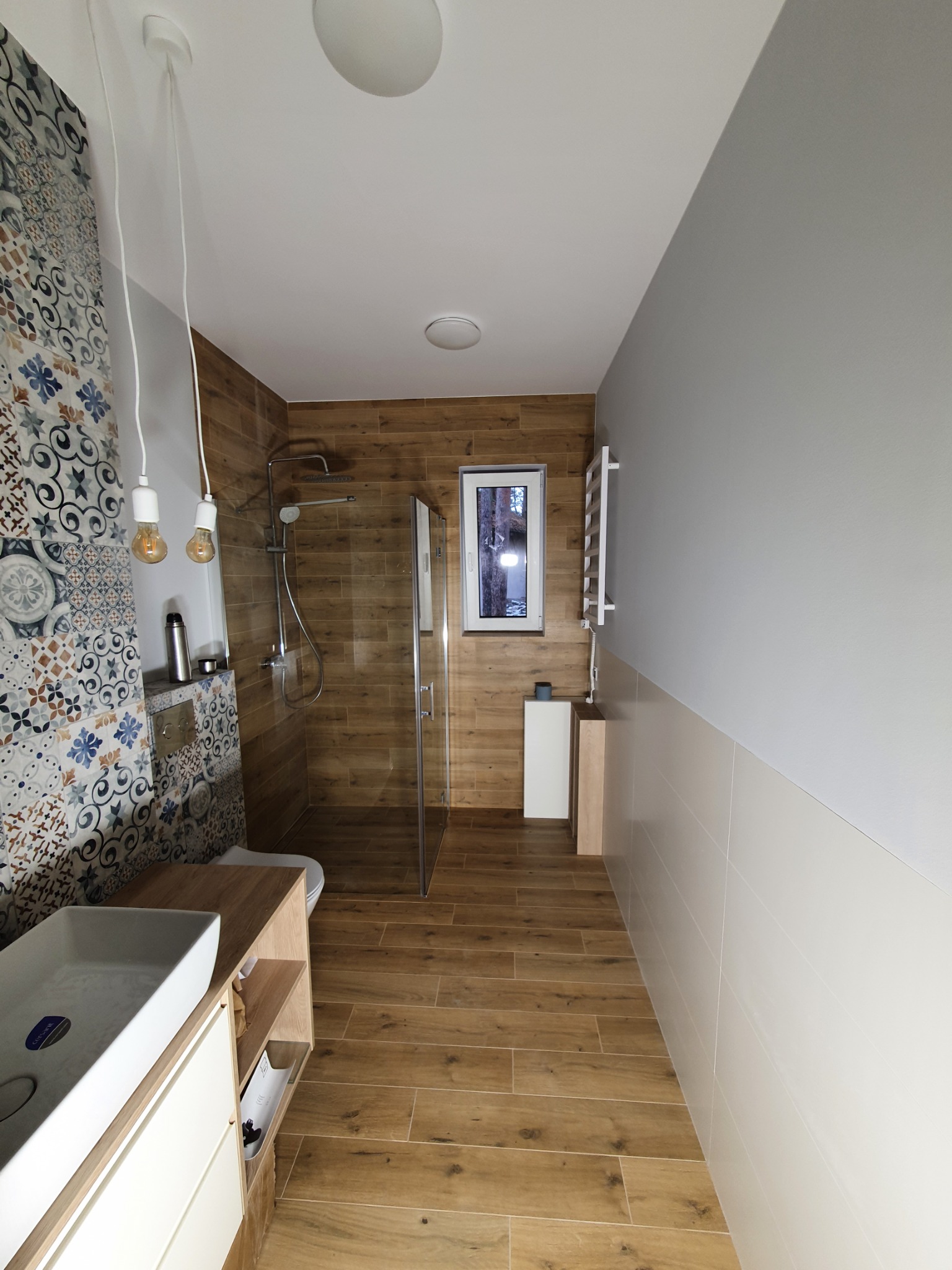 Nowoczesna łazienka z kabiną prysznicową typu walk-in, drewnopodobnymi płytkami na podłodze i ścianach, umywalką nablatową na szafce z jasnego drewna oraz dekoracyjną ścianą z mozaiki wzorzystej.