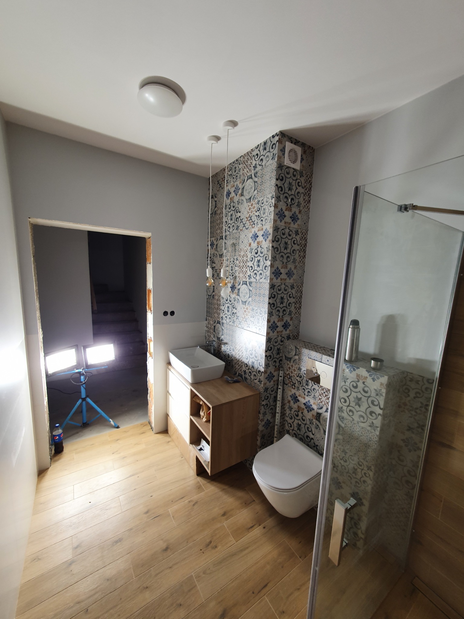 Wnętrze łazienki w trakcie remontu, widoczne płytki z wzorem marokańskim, drewnopodobna podłoga, umywalka z szafką, toaleta podwieszana oraz kabina prysznicowa; w tle widoczny korytarz...