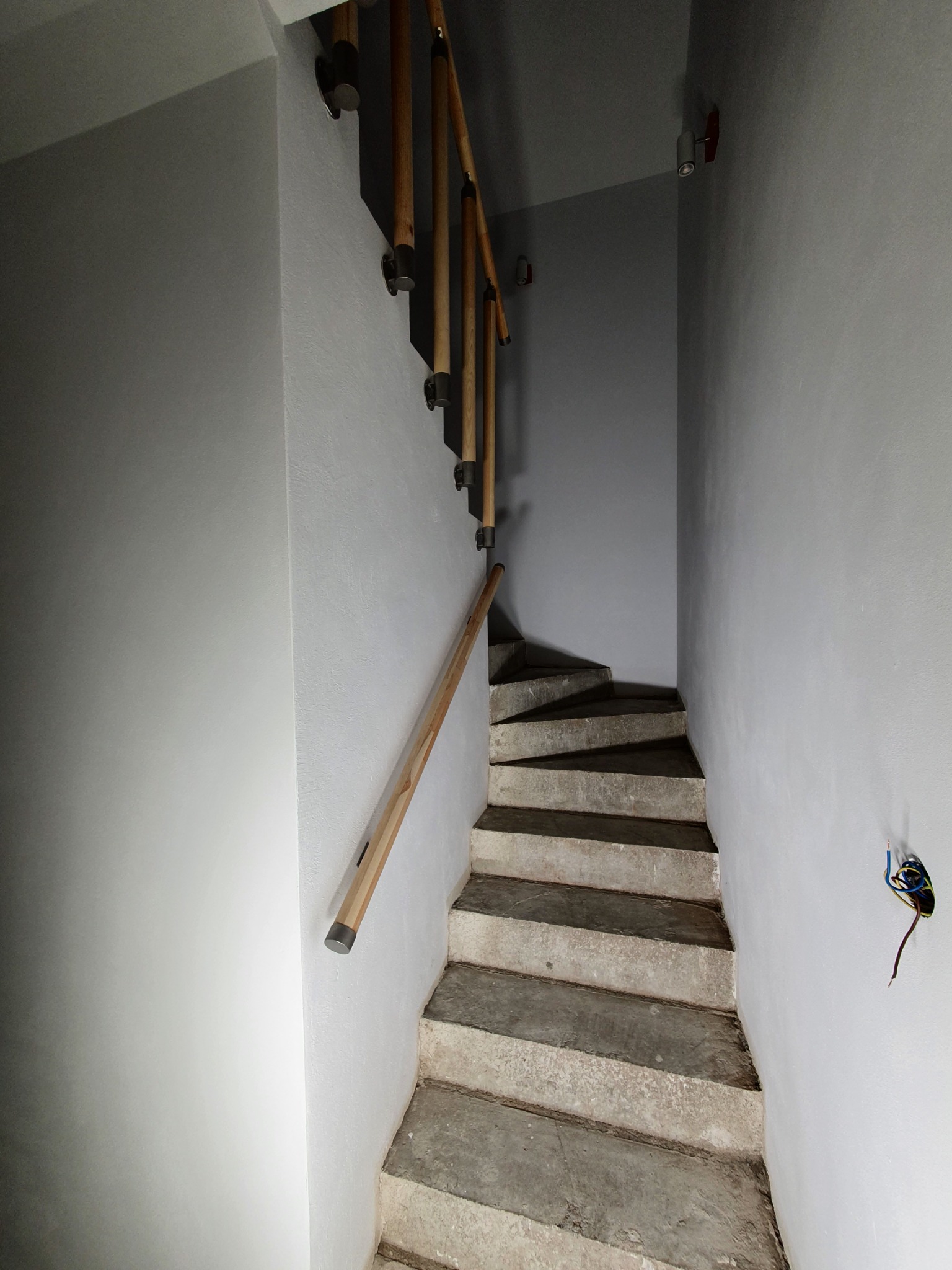 Klatka schodowa w szarych tonacjach z drewnianą balustradą, surowe betonowe schody, wystające przewody elektryczne na ścianie.