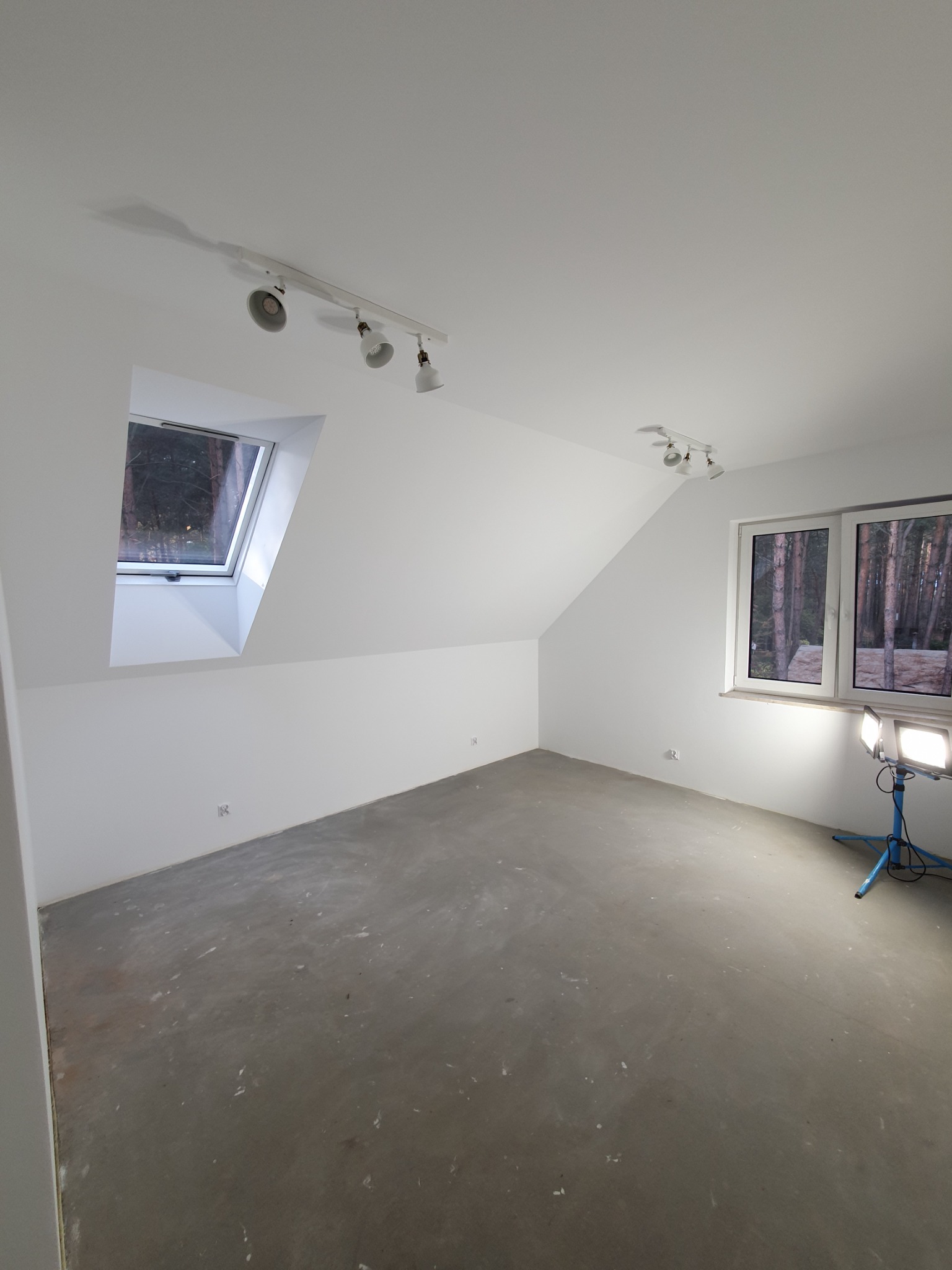 Pokój na poddaszu w Radzyminie przed malowaniem, z widocznym oknem dachowym, oknem standardowym oraz lampą budowlaną na trójnogu