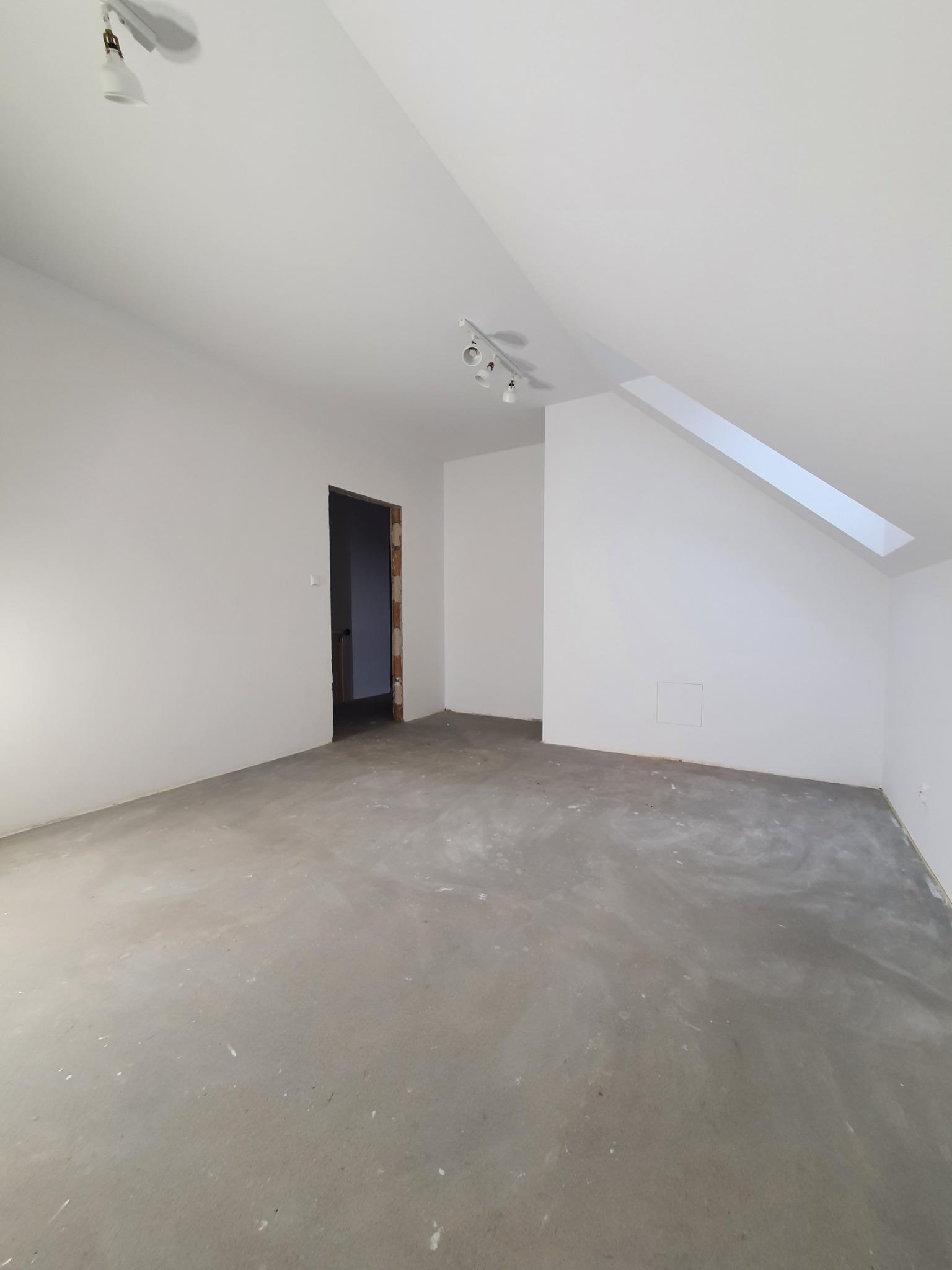 Pusty pokój na poddaszu z surową podłogą, świeżo pomalowanymi białymi ścianami i oknem dachowym, przygotowany do dalszych prac wykończeniowych.