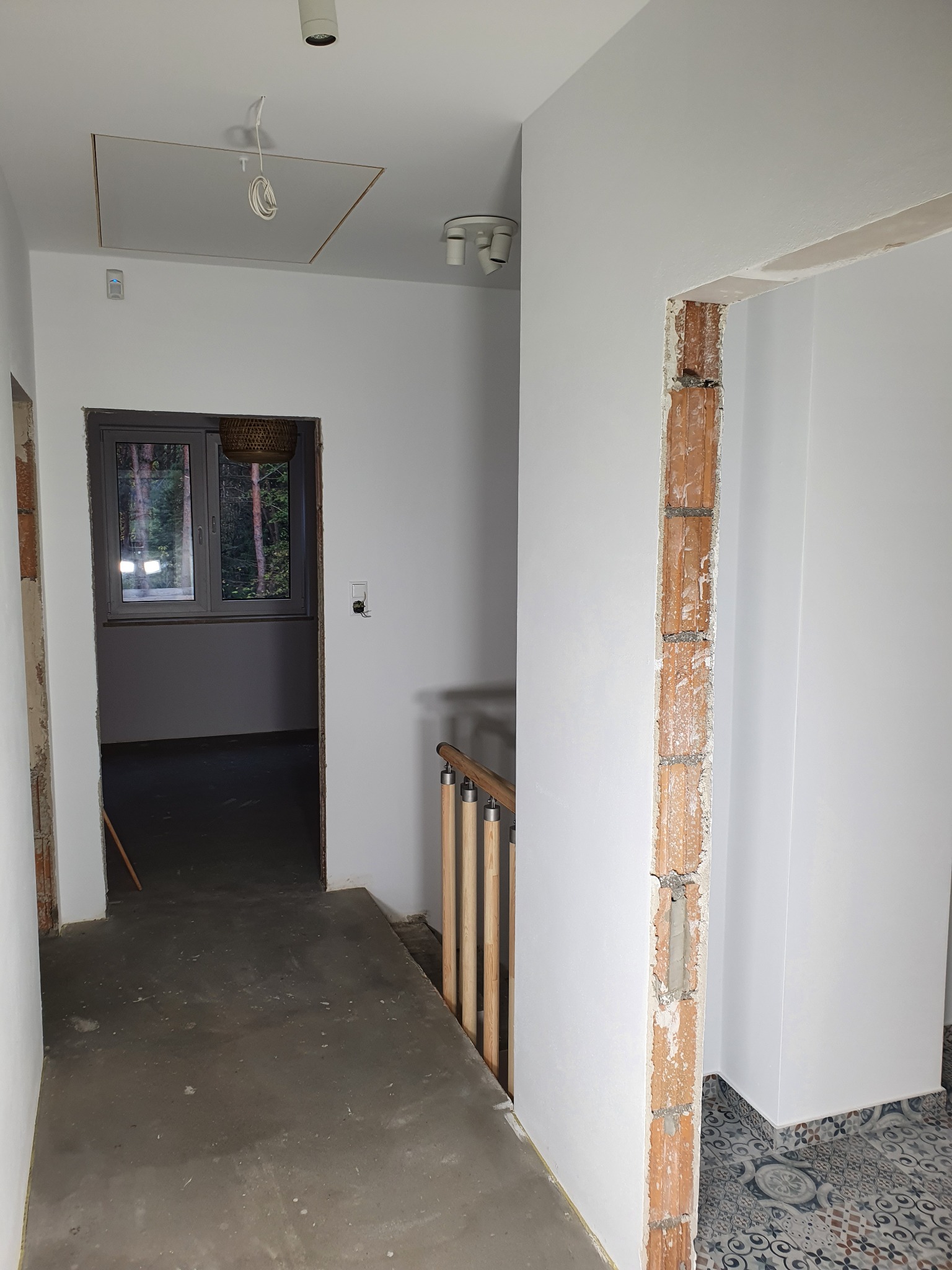 Wnętrze w trakcie prac wykończeniowych: białe ściany, surowy otwór drzwiowy z odsłoniętą cegłą, widoczne instalacje elektryczne na suficie, fragment drewnianej balustrady schodów, okno z widokiem...