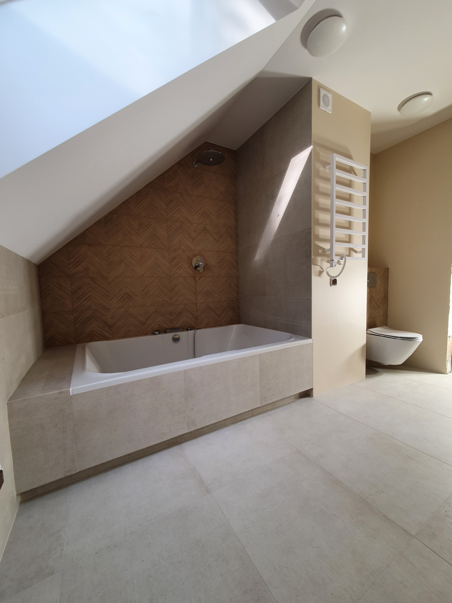 Jasna łazienka na poddaszu z wanną obudowaną płytkami w odcieniach beżu, mozaikową ścianą prysznicową w jodełkę i podwieszaną toaletą, oświetlona światłem z okna dachowego.