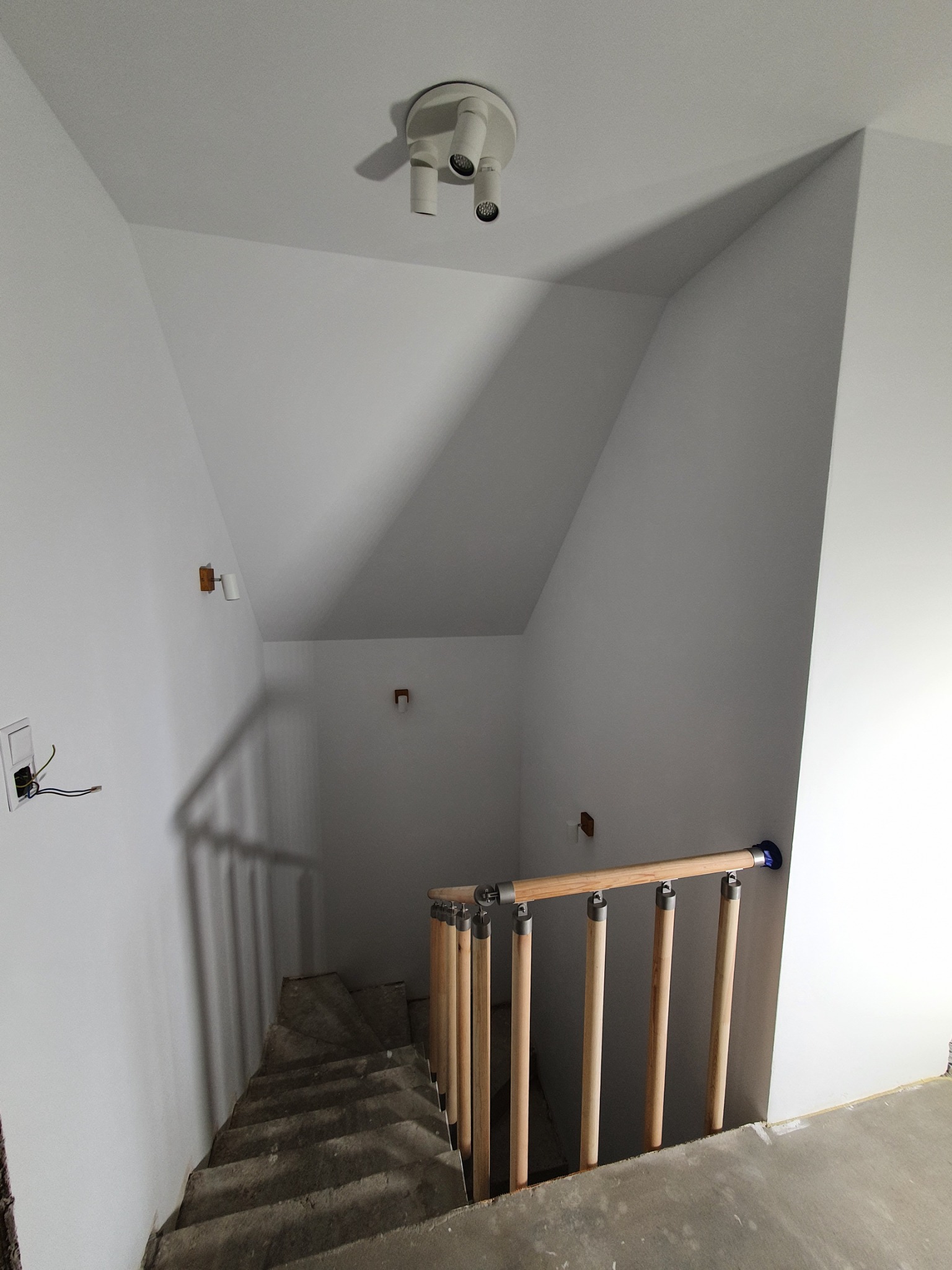 Klatka schodowa w trakcie wykańczania: surowe schody betonowe, białe ściany z widocznymi instalacjami elektrycznymi, drewniana balustrada z metalowymi elementami, oświetlenie sufitowe i ścienne.