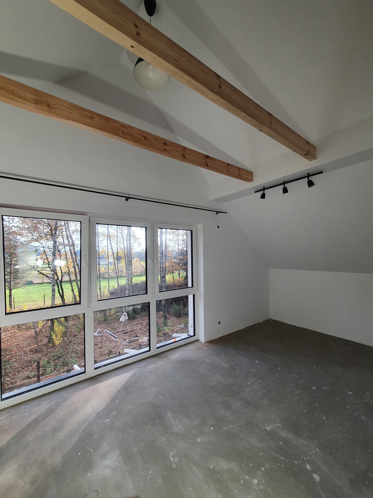 Poddasze w trakcie wykańczania: surowa wylewka na podłodze, białe ściany, drewniane belki stropowe, okno z widokiem na jesienny krajobraz i zamontowane czarne reflektory sufitowe.