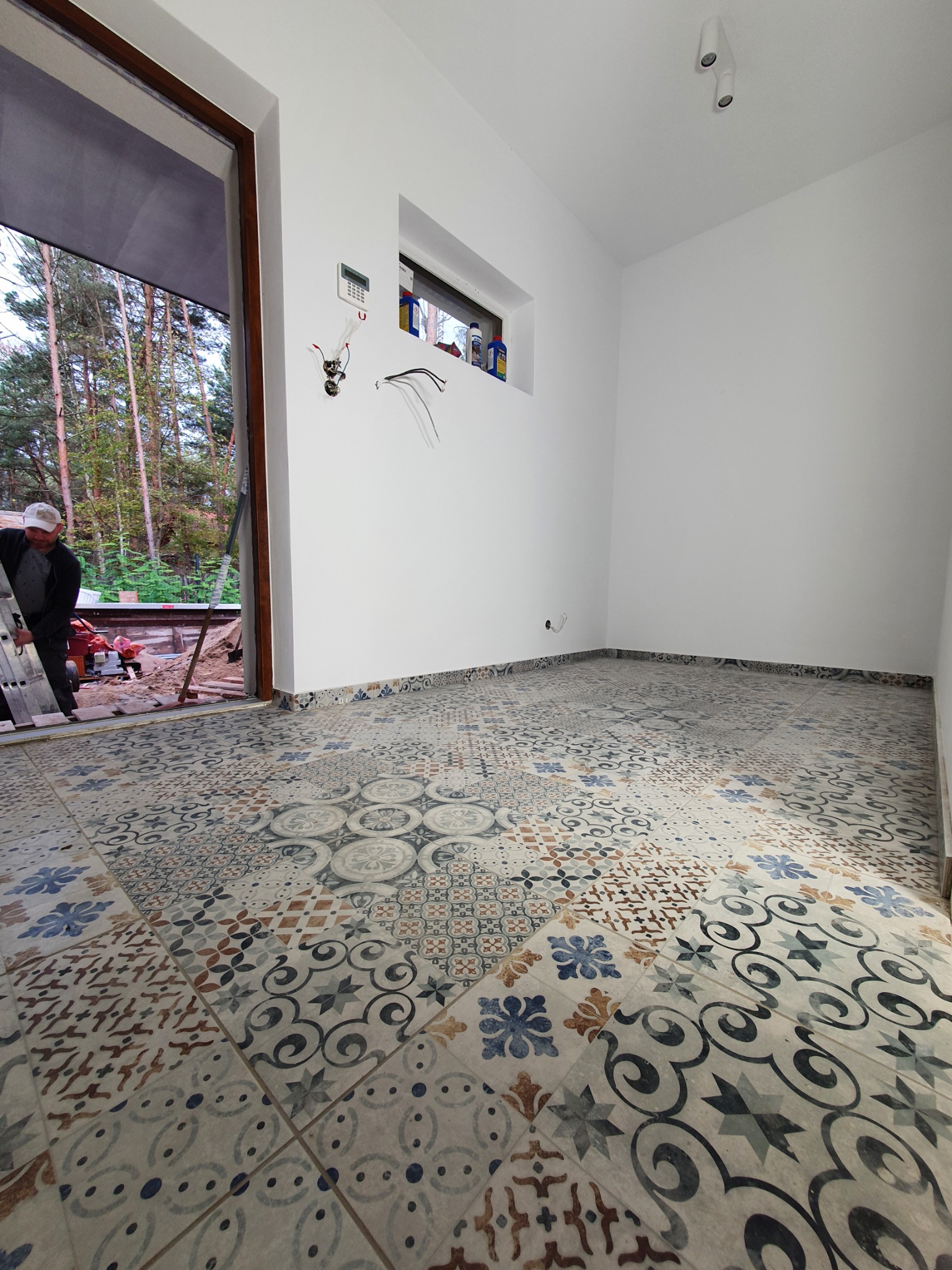 Wnętrze w trakcie prac wykończeniowych: świeżo ułożona, wzorzysta podłoga z płytek ceramicznych, białe ściany z zamontowanym oknem i wystającymi przewodami elektrycznymi, widoczny fragment drabiny...