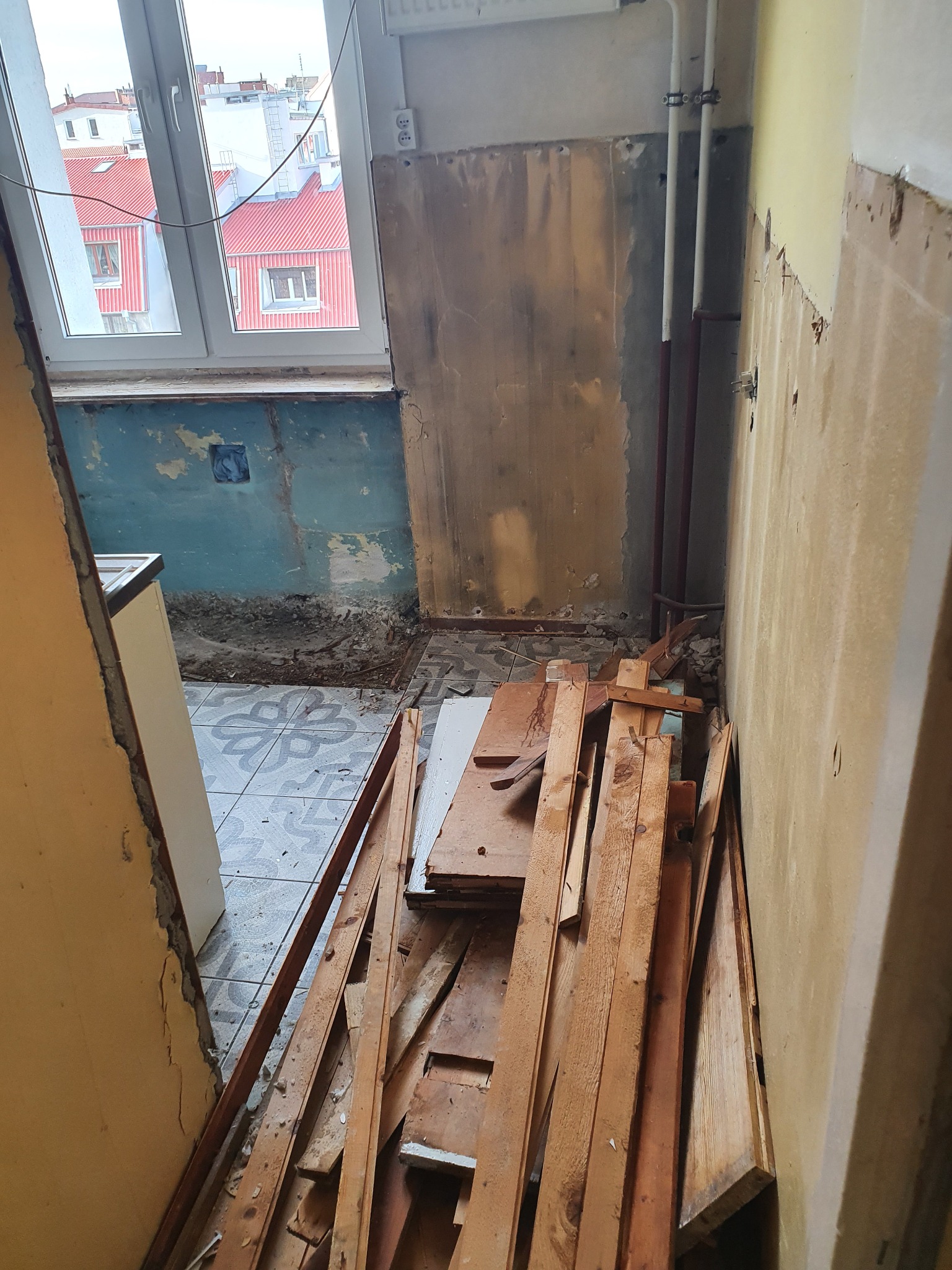 Wnętrze w trakcie demontażu, widoczne stare, niebieskie ściany z resztkami tapety, odrapane tynki, fragmenty drewnianych listew i desek, oraz odsłonięte rury grzewcze przy ścianie.