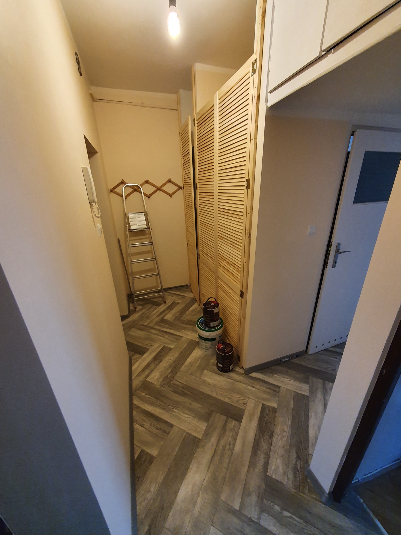 Wnętrze korytarza w trakcie prac, widoczna drabina oparta o ścianę, puszki z farbą przy drewnianej zabudowie z żaluzjowymi drzwiami, podłoga wyłożona panelami w jodełkę.