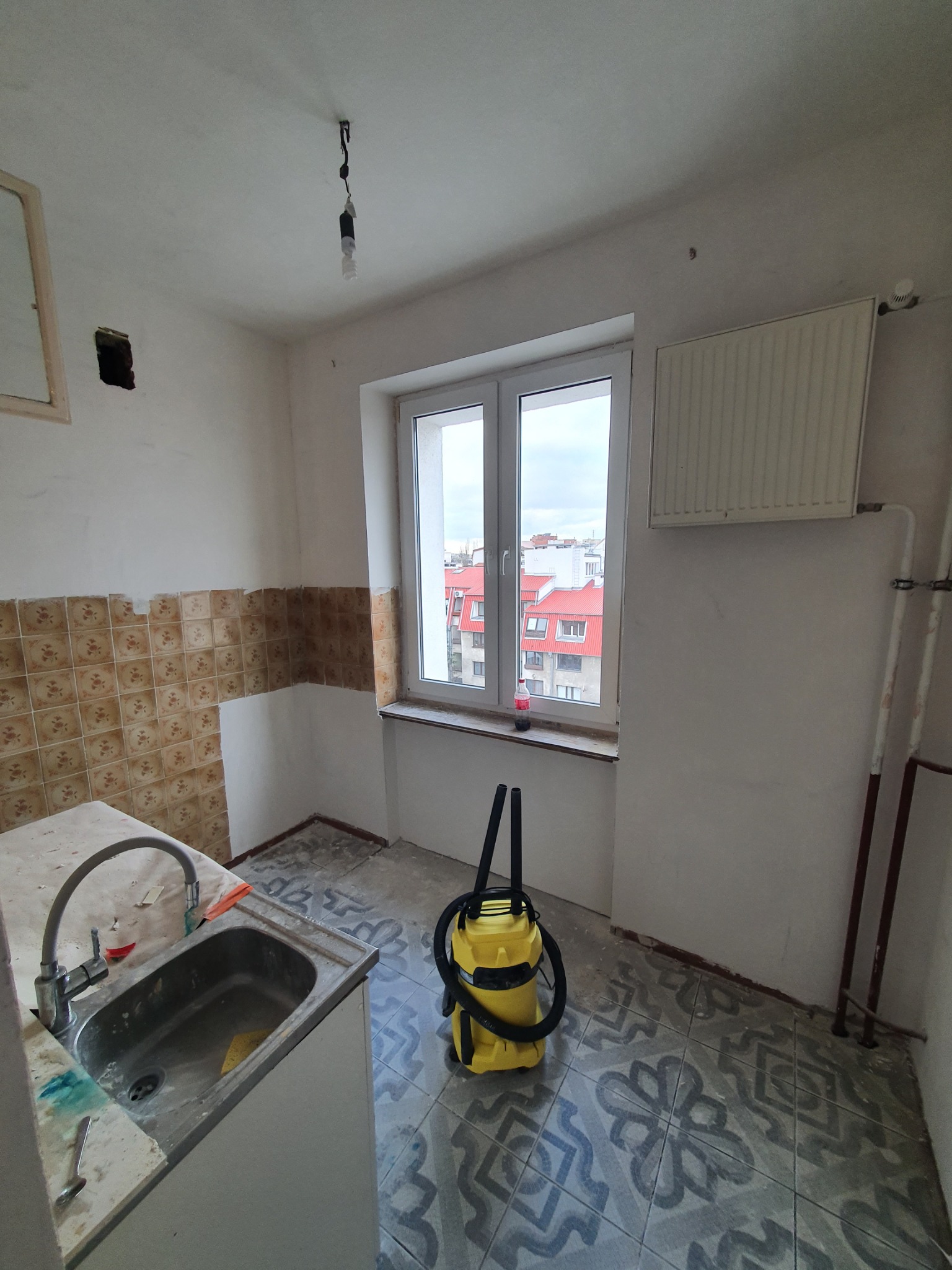 Wnętrze kuchni w trakcie demontażu płytek ściennych i podłogowych, widoczny żółty odkurzacz przemysłowy, okno z widokiem na budynki z czerwoną dachówką i odsłonięte przewody elektryczne.