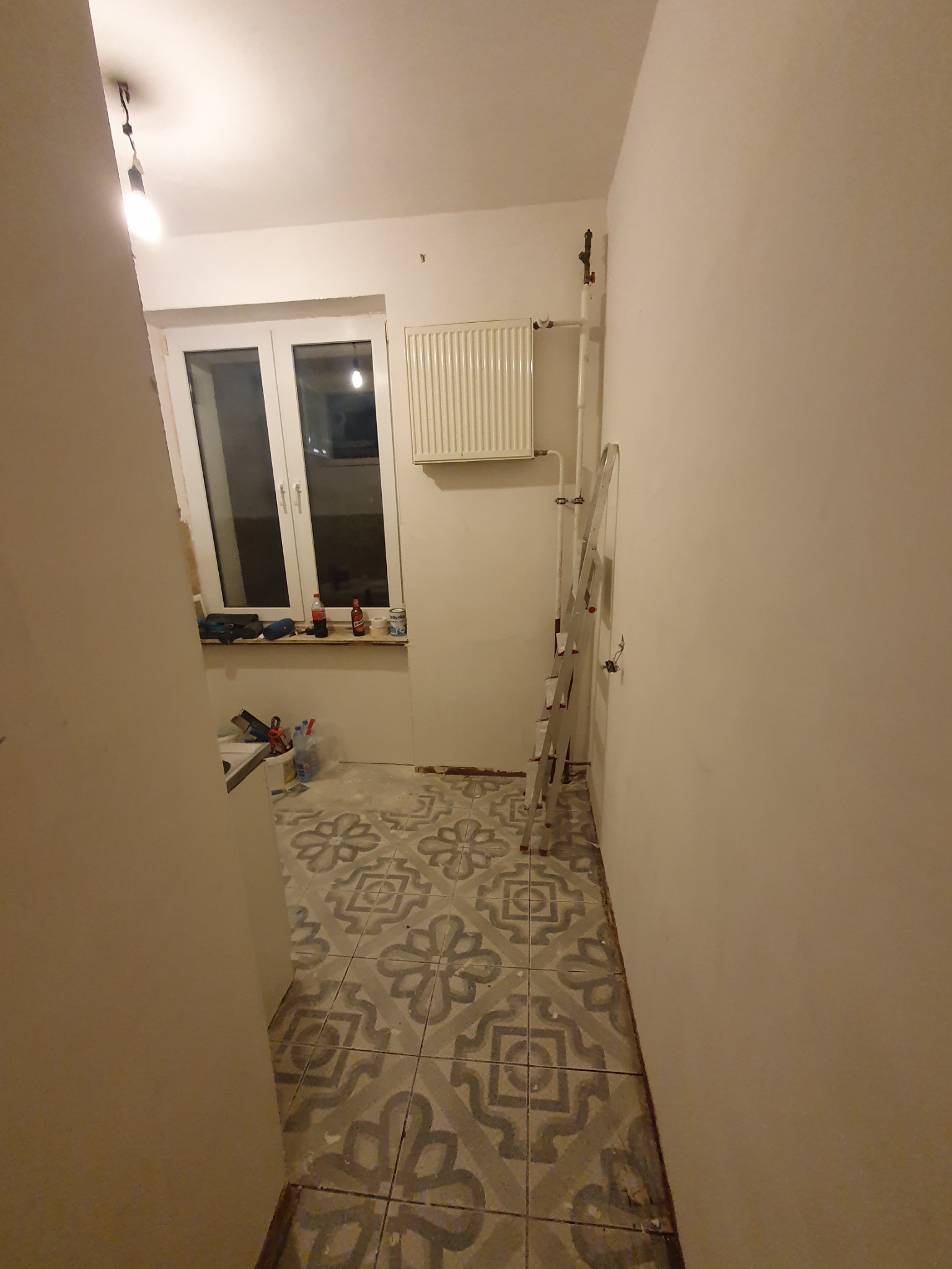 Wnętrze w trakcie remontu: widoczne płytki podłogowe z wzorem, drabina oparta o ścianę, grzejnik, okno z narzędziami i materiałami budowlanymi na parapecie, surowy stan ścian.