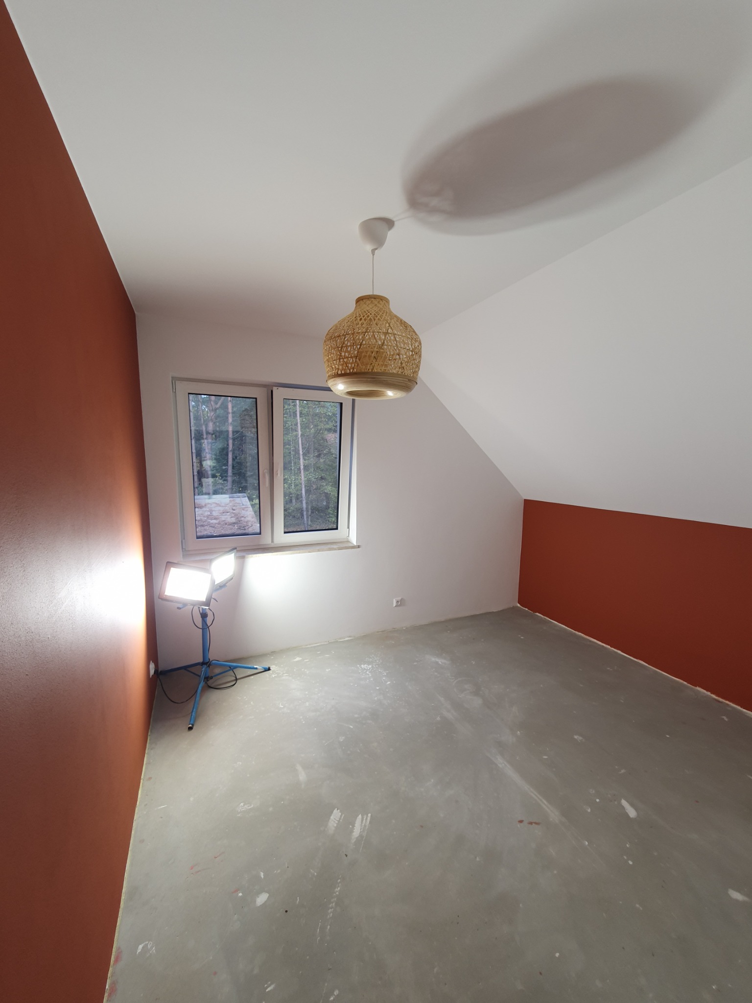 Pokój w trakcie malowania z lampą roboczą na trójnogu, częściowo pomalowane ściany w kolorze terakoty, surowa podłoga, okno z widokiem na las i pleciona lampa wisząca.