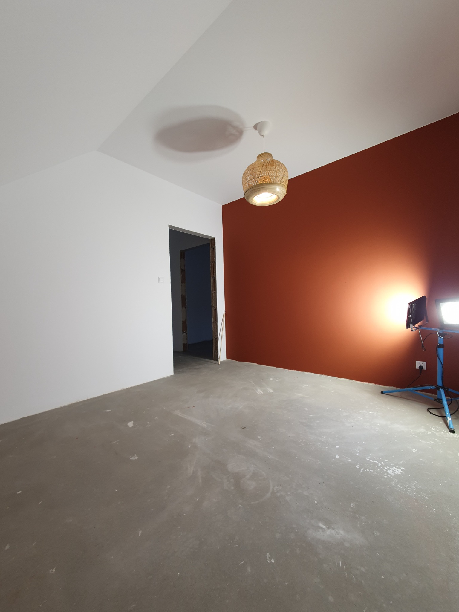 Pokój w trakcie remontu: jedna ściana pomalowana na terakotowy kolor, druga biała, surowa podłoga, widoczna lampa wisząca z abażurem z plecionki i lampa budowlana na trójnogu.