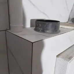 Półka obłożona białymi płytkami imitującymi marmur z widocznym czarnym pierścieniem uszczelniającym i fragmentem białej rury kanalizacyjnej w rogu pomieszczenia.