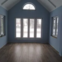 Wnętrze pokoju z drewnopodobną podłogą, niebieskimi ścianami i białym, skośnym sufitem z oknem łukowym na szczycie.