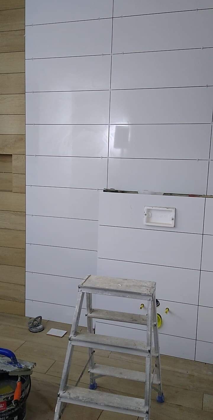 Ściana w trakcie układania białych, prostokątnych płytek z widocznymi fugami, obok fragment ściany wykończonej drewnopodobnymi panelami, aluminiowa drabina stojąca przed ścianą z przygotowanymi...