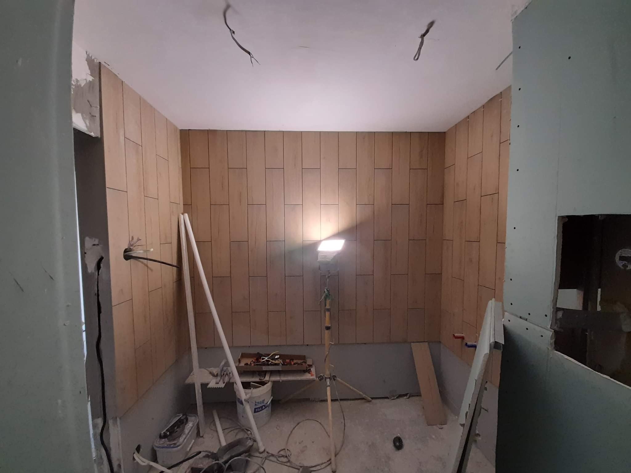 Wnętrze w trakcie prac wykończeniowych: ściany częściowo pokryte panelami imitującymi drewno, widoczna instalacja elektryczna, narzędzia pracy i lampa na statywie.