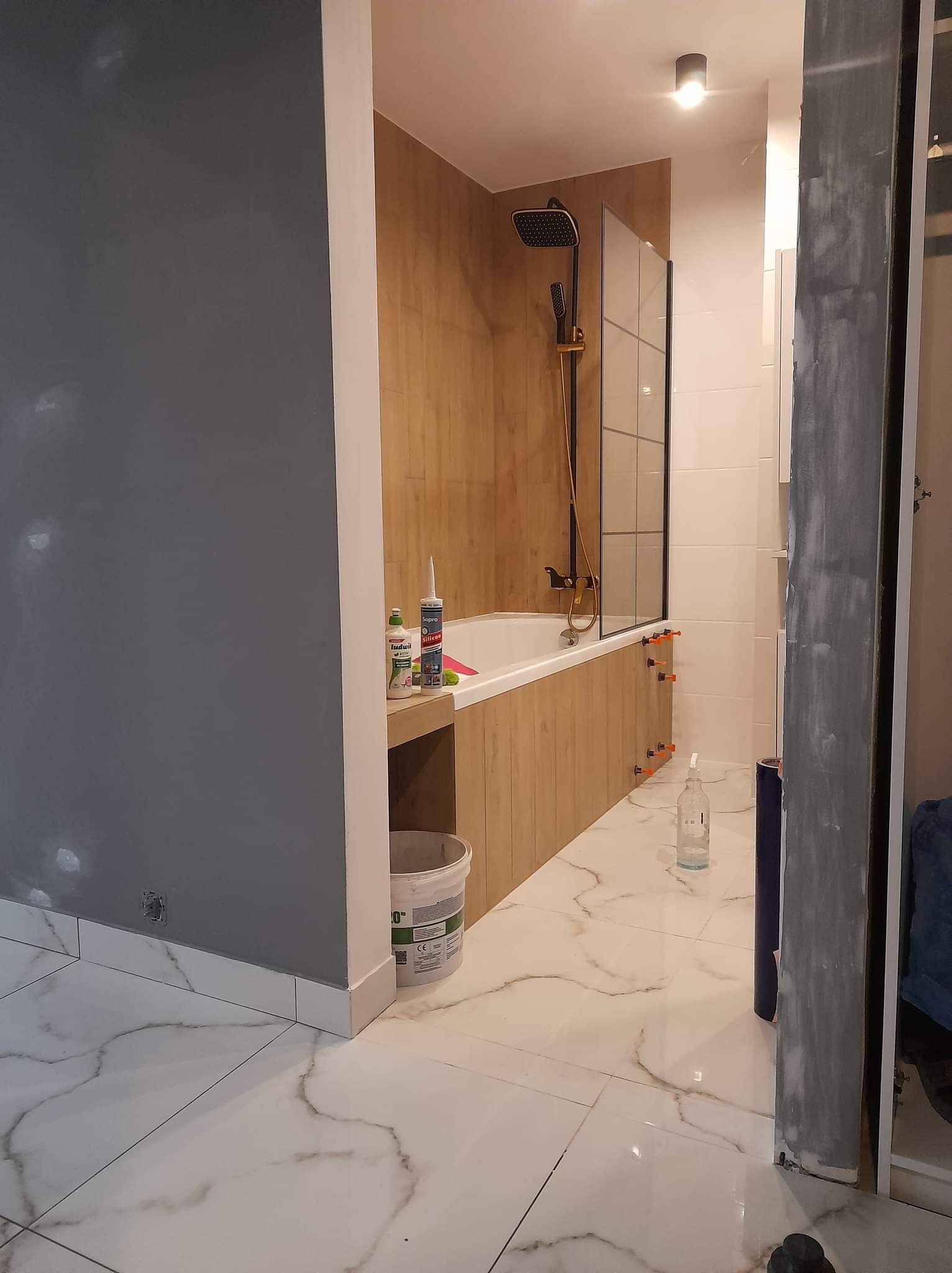 Nowoczesna łazienka w trakcie wykończenia: drewnopodobna zabudowa wanny z czarną armaturą, białe marmurowe płytki podłogowe, szara ściana i narzędzia pracy.