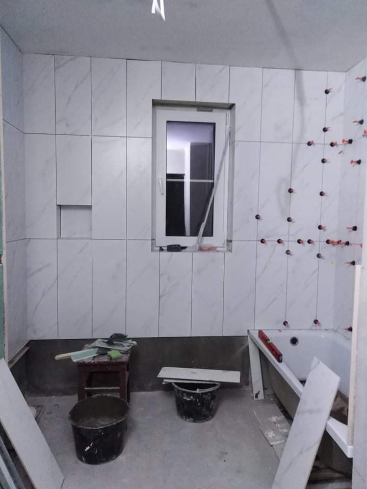 Łazienka w trakcie remontu: białe płytki imitujące marmur układane na ścianach, widoczne krzyżyki dystansowe, okno z białymi ramami, narzędzia i wiaderka na podłodze.