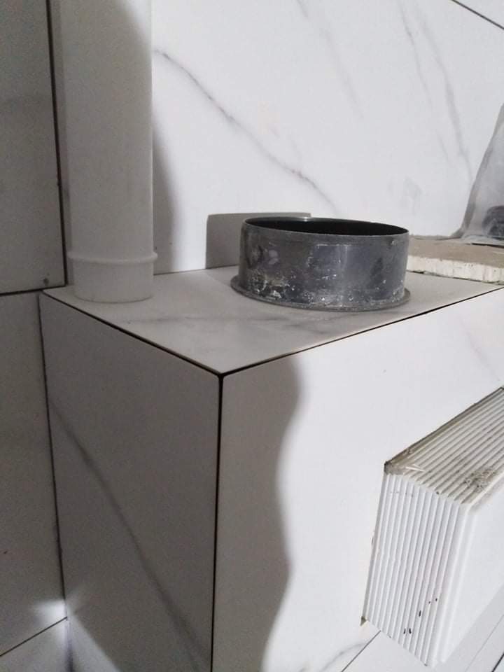 Półka obłożona białymi płytkami imitującymi marmur z widocznym czarnym pierścieniem uszczelniającym i fragmentem białej rury kanalizacyjnej w rogu pomieszczenia.