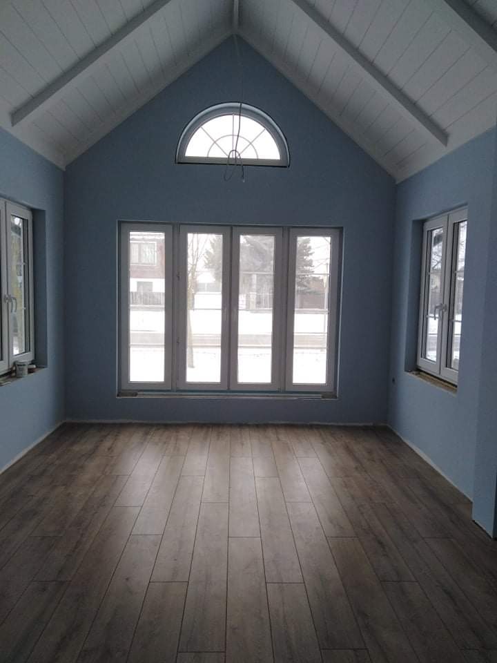 Wnętrze pokoju z drewnopodobną podłogą, niebieskimi ścianami i białym, skośnym sufitem z oknem łukowym na szczycie.