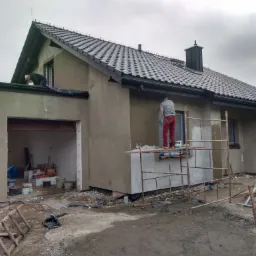 Dwóch pracowników na rusztowaniach podczas prac elewacyjnych przy domu jednorodzinnym, elewacja w trakcie tynkowania, widoczny garaż, niebieskie niebo w tle.