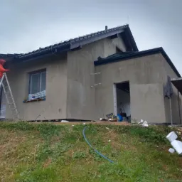 Pracownik w pomarańczowej kurtce na drabinie maluje fragment elewacji domu jednorodzinnego w trakcie ocieplania, widoczne styropianowe płyty na trawie i narzędzia.