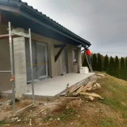 Pracownik w pomarańczowej kurtce na drabinie przy domu z elewacją w trakcie prac, widoczne rusztowanie, okna zabezpieczone folią, tło z rzędem wysokich tuj.