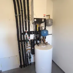 Nowoczesna kotłownia z białym zasobnikiem ciepłej wody, piecem gazowym Viessmann i rozbudowaną instalacją hydrauliczną z czarnymi rurami izolowanymi termicznie, zamontowana w narożniku...