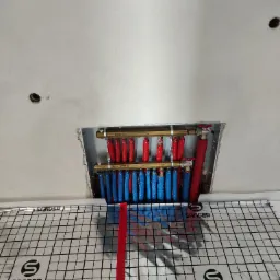 Rozdzielacz instalacji grzewczej z rurami w izolacji czerwonej i niebieskiej, widoczny przez otwór w suficie, z folią aluminiową z nadrukiem siatki i logo na podłodze.