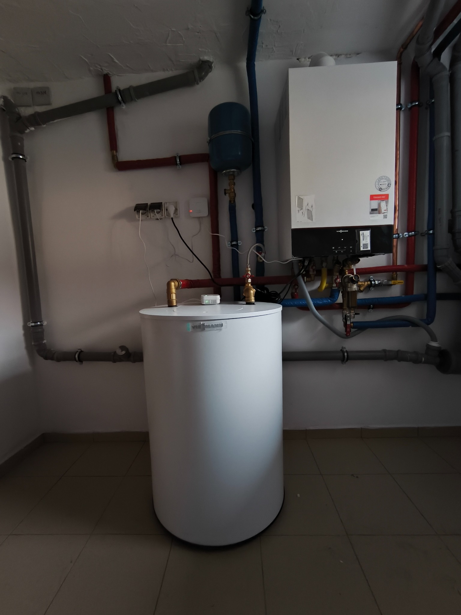 Biały zasobnik ciepłej wody Viessmann z podłączeniami, rurami i piecem grzewczym na ścianie w pomieszczeniu technicznym, widok z dołu.