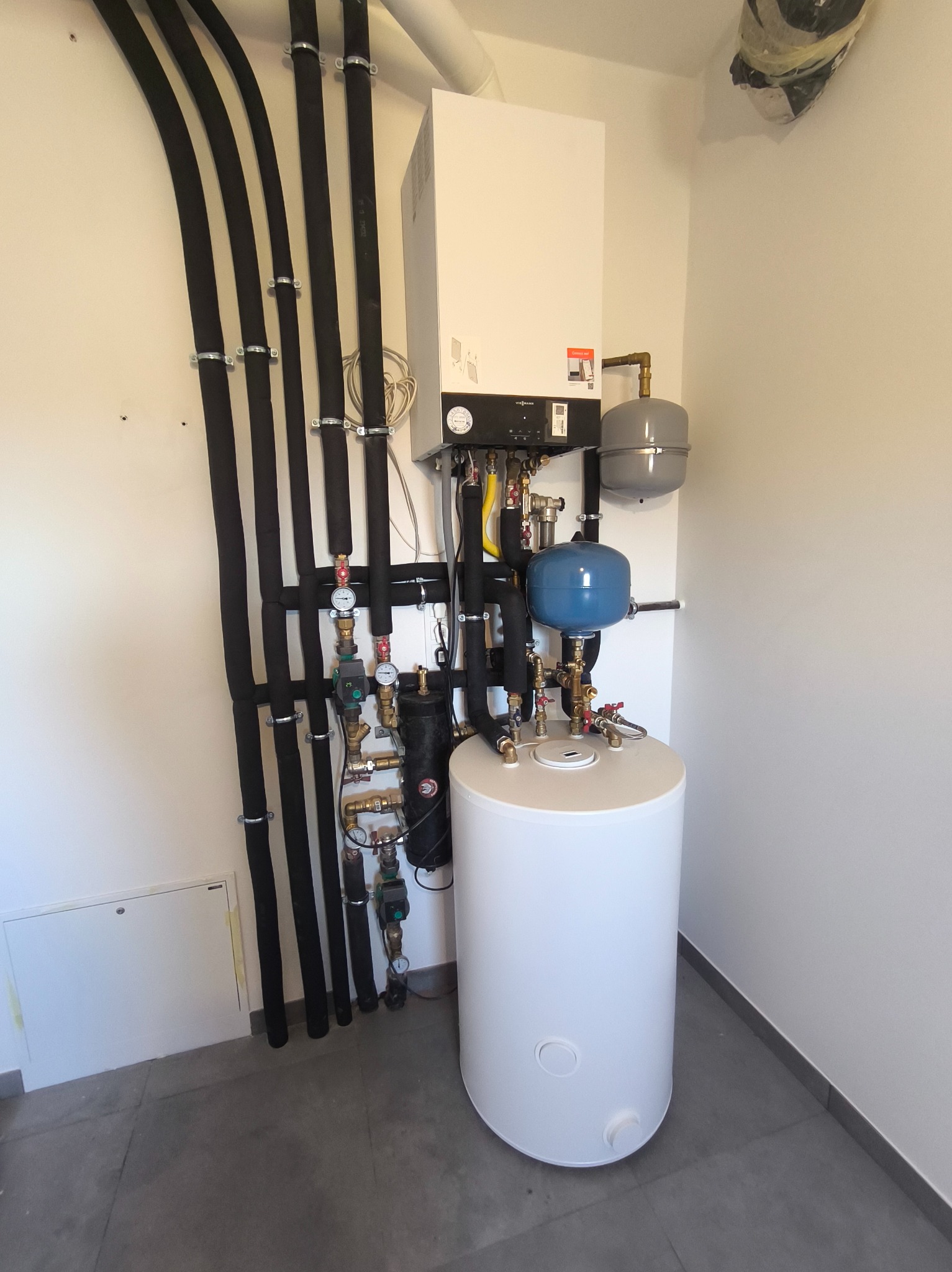 Nowoczesna kotłownia z białym zasobnikiem ciepłej wody, piecem gazowym Viessmann i rozbudowaną instalacją hydrauliczną z czarnymi rurami izolowanymi termicznie, zamontowana w narożniku...