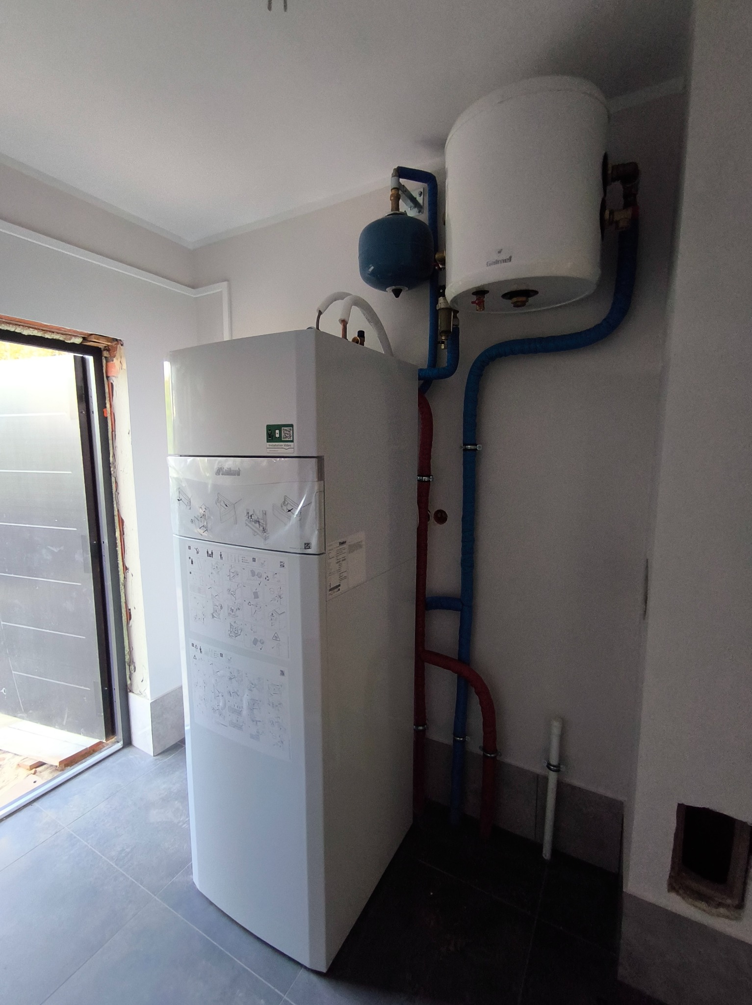 Biały piec grzewczy Vaillant z niebieskimi i czerwonymi rurami, zbiornikiem wyrównawczym i bojlerem zamontowany w pomieszczeniu z szarą podłogą i otwartymi drzwiami.