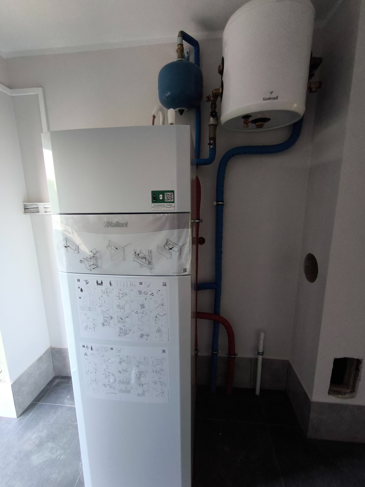 Nowoczesny, biały piec gazowy Vaillant w folii ochronnej, podłączony do instalacji z niebieskimi i czerwonymi rurami, obok zbiornik Galmet, widoczne instrukcje montażu na obudowie.