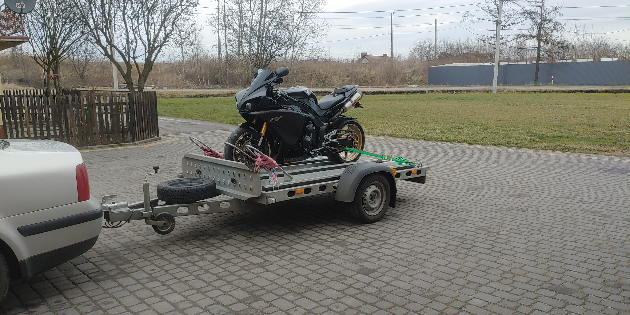 Czarny motocykl Yamaha R1 zabezpieczony pasami na metalowej przyczepie, podpiętej do srebrnego samochodu osobowego na tle brukowanej nawierzchni i krajobrazu z drzewami bez liści.