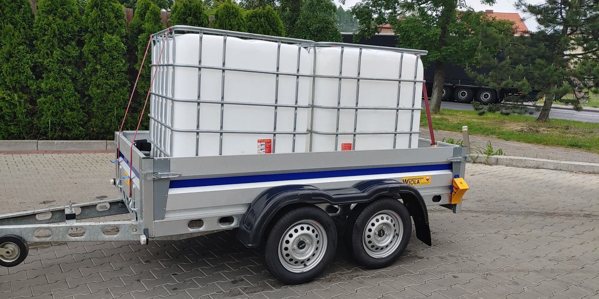 Przyczepa samochodowa z dwoma białymi zbiornikami IBC zabezpieczonymi pasami transportowymi, widok z boku na tle zieleni i drogi.