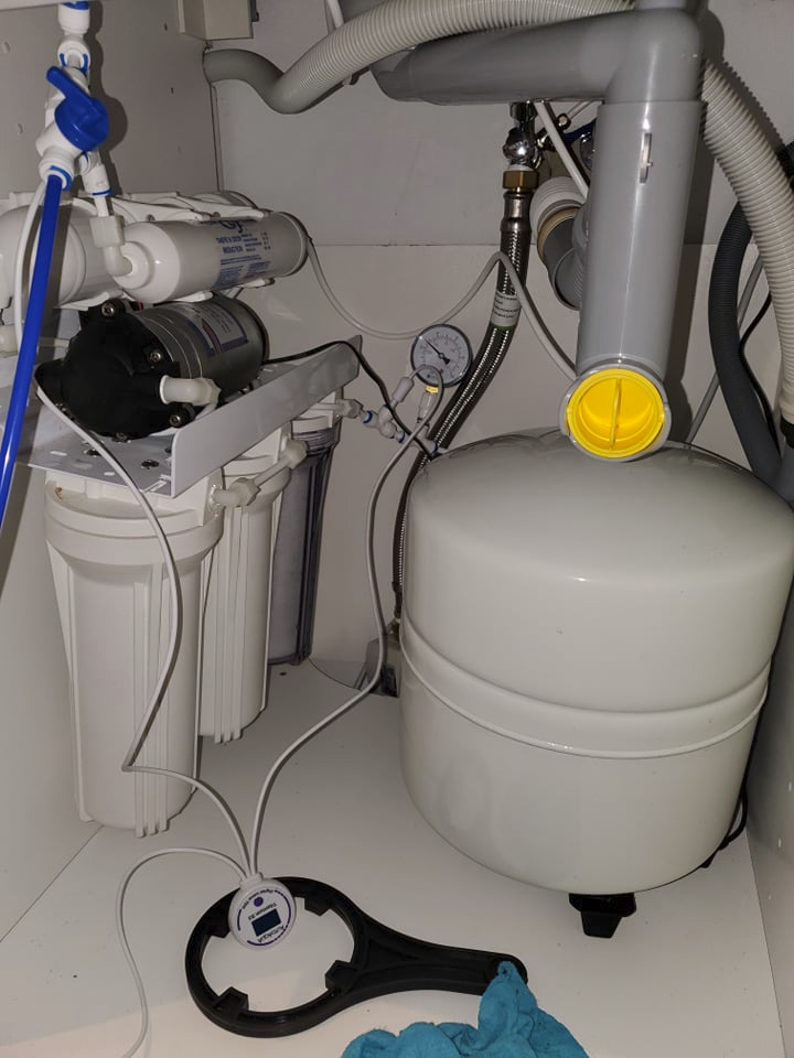 Zamontowany system odwróconej osmozy pod zlewem, widoczne filtry, zbiornik ciśnieniowy, manometr i klucz do obudowy filtra. Niebieski zawór odcinający wodę. Białe wnętrze szafki.